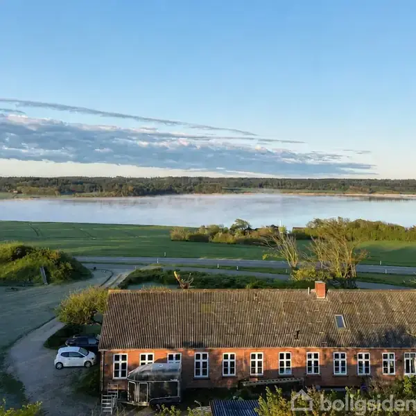 En rød murstensbygning med skråtag står stille i et landligt område. Foran bygningen ses grønne marker, og i baggrunden strækker en stille sø sig under en blå himmel.