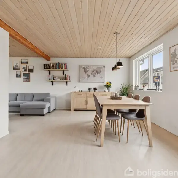 Et lyst og rummeligt rum med spisebord og stole i forgrunden. En grå sofa står ved siden af et skænk, bøger og vægkunst. Store vinduer giver naturligt lys.