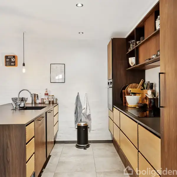Køkken med træskabsfacader og mørk bordplade, arrangeret med stilfulde lamper og køkkenartikler. Lysegrå gulvfliser og minimalistisk indretning skaber en moderne atmosfære. En tavle viser datoen den 10.