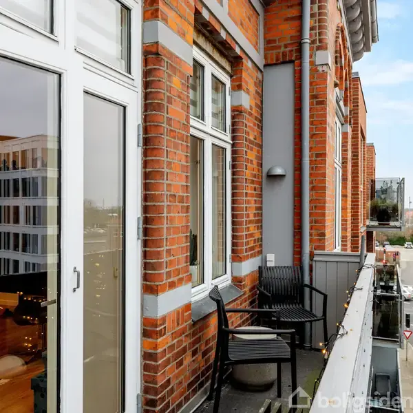 En altan med to sorte stole står mod en facade af røde mursten. Gaden nedenfor vises med biler og bygninger i baggrunden under en blå himmel.