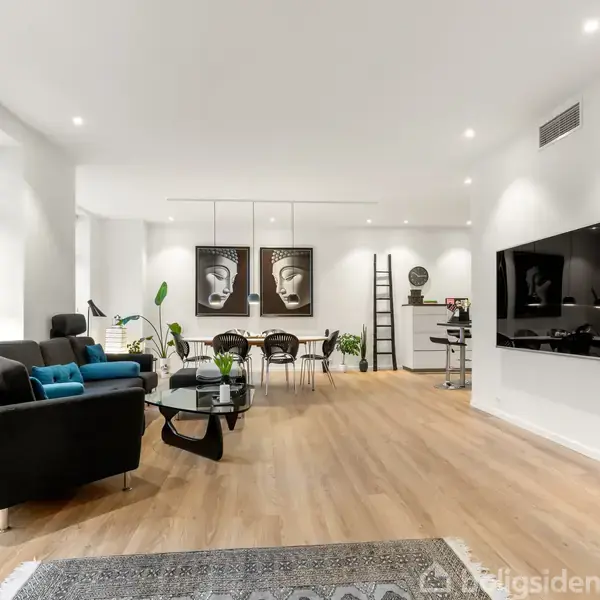 En moderne stue med sorte lædersofaer, hvor der står grønne planter rundt omkring. Væggene er hvide med kunst på dem, og der er et stort TV monteret.