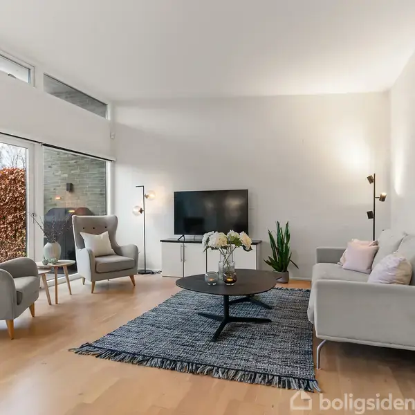 En stue indeholder en sofa og to lænestole der omkranser et lavt sofabord med blomster. Et tæppe dækker trægulvet. En fladskærms-TV står foran store vinduer med udsigt til en have.