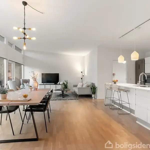 Stue med spisebord og stole til venstre, sofaområde med tv midt i rummet, og et moderne køkken til højre. Store vinduer slipper lys ind og viser en terrasse udenfor.