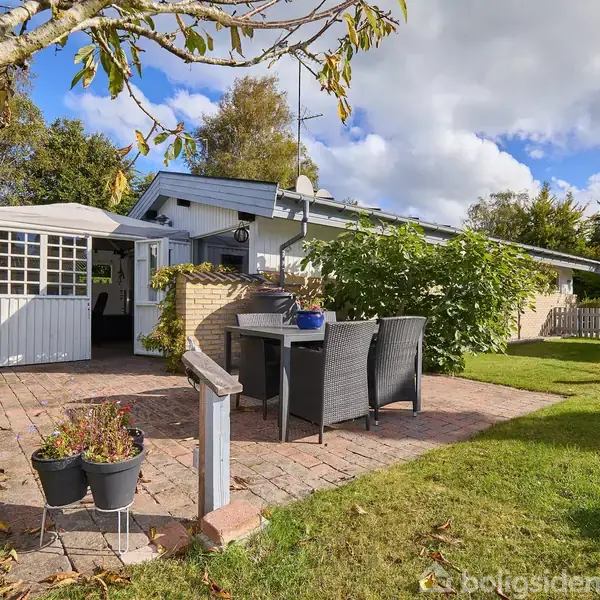 Et hus står omgivet af en have med en flisebelagt terrasse. Terrassen har et bord med stole, planter og en åben dør til huset. Skyer ses på himlen.