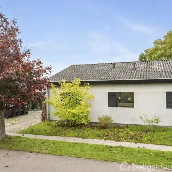 Et hvidt hus med grå tagsten står stille i et boligområde. Foran er en grøn græsplæne, en flisebelagt sti, og et træ med røde blade.
