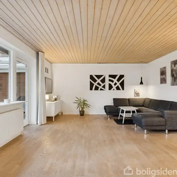 Sofa står i en lys stue med trægulv, ved siden af et sofabord og en lampe. Store vinduer åbner op mod en terrasse. Vægkunst og planter pynter rummet.