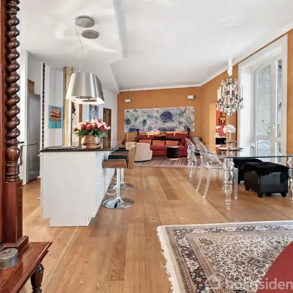 Et moderne køkken-alrum med trægulv, hvidt køkkenø, og krystallysekroner. Plastikstole og et maleri pryder rummet, som har lyse vægge og store vinduer. Møblerne er stilfulde.