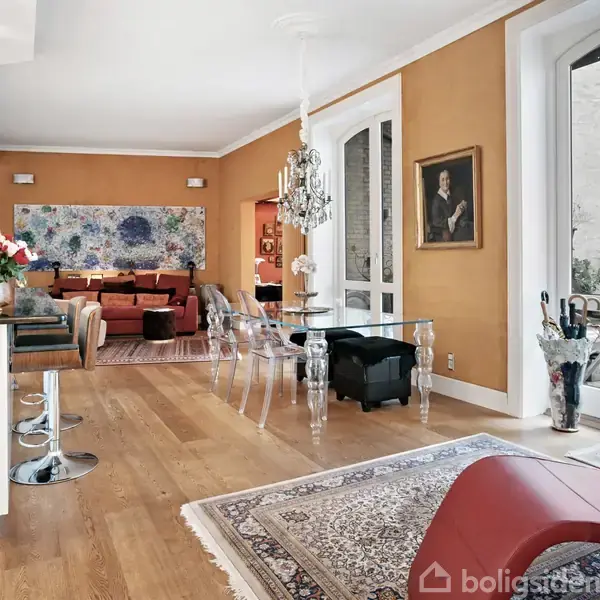 Et moderne, stilfuldt rum med trægulv og orange vægge. En glasbord omgivet af stole, står centralt. En stor sofa er i baggrunden, tæt på kunstværk. Store vinduer til højre. En buket med blomster og lysekrone tilføjer elegance. En portrætmaleri hænger på væggen.