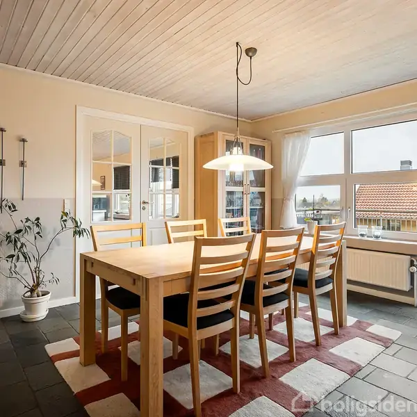 Et spisebord med syv stole står på et lysebrunt og rødt tæppe. En lampe hænger over bordet, og store vinduer giver udsigt til et boligområde med røde tage.