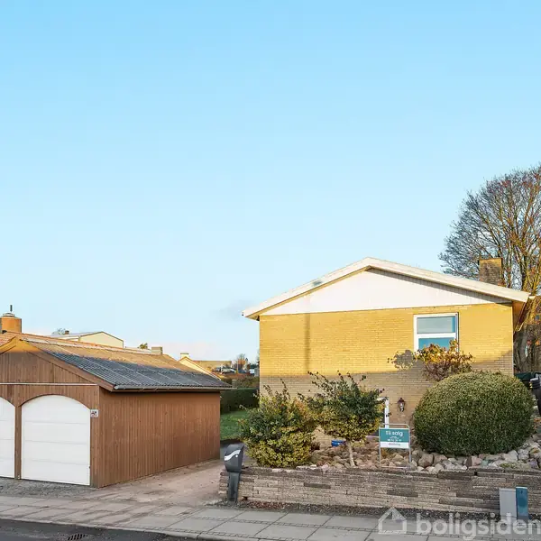 Et gult hus med fladt tag står bag en stenet have. Ved siden af er en garage med brune porte. Omgivet af træer og nabohuse under en klar himmel.