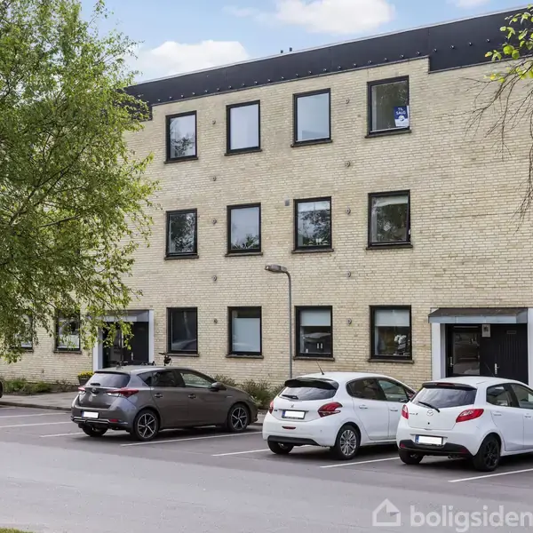 Bygning med gule mursten og tre etager, parkerede biler foran, omgivet af træer og fortov.