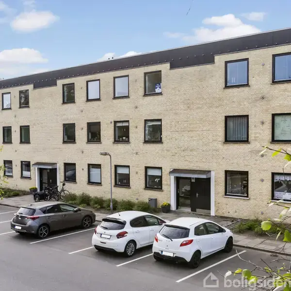 Et tre-etagers gult murstensbyggeri med sort tag står i en parkeringsplads, hvor fem biler er parkeret foran. Grønne træer og blå himmel danner baggrunden.