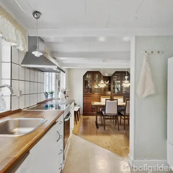 Køkken med vask og træbordplade, der strækker sig mod en spisestue med mørke træskabe og spisebord. Store vinduer til venstre lader lys ind, hvilket lyser rummet op.