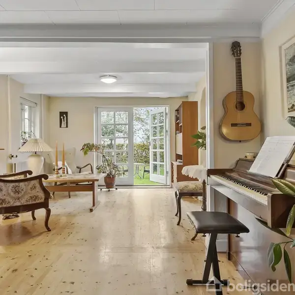 Et værelse indeholder et klaver med nodeside, omgivet af planter. En akustisk guitar hænger på væggen. I rummet ses også stole og et bord. En åben dør fører ud til en have.