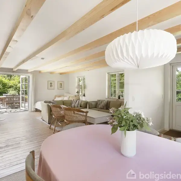 Spisebord med lyserødt dug står centralt i et lyst opholdsrum. Over bordet hænger en hvid, foldet lampe. I baggrunden er en sofagruppe og en åben dobbeltdør til en træterrasse.