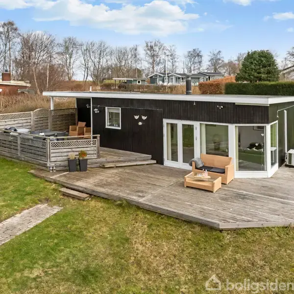 Et sort modernistisk sommerhus med hvide vinduer og fladt tag står på en træterrasse. Omringet af græsplæne og hække, med sparsomt træbeklædt landskab i baggrunden.