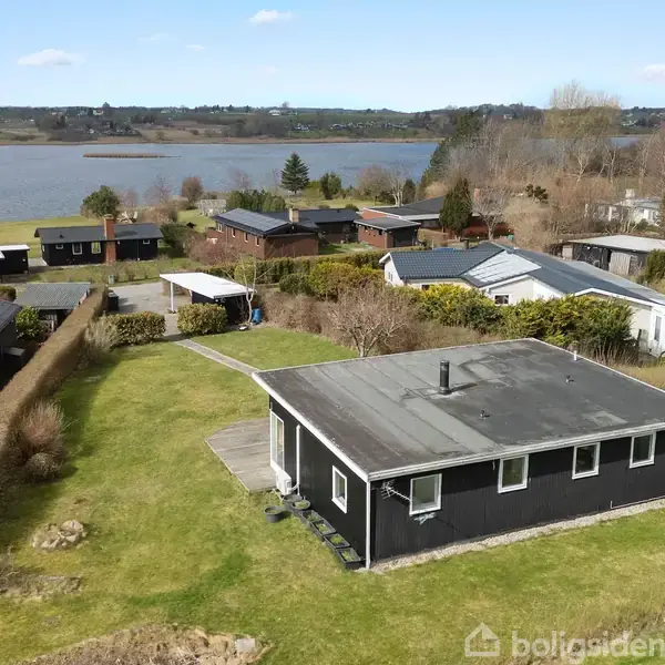 Et sort, firkantet hus med fladt tag står stille i en grøn have. Det omgivende landskab inkluderer flere huse, træer og en stor sø i baggrunden.