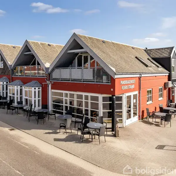 Rødt hus med skilt "Pandekagehuset Vejers Strand," omgivet af udendørs borde og stole. Solrigt vejr med blå himmel, beliggende ved en gade med fortov og grønne områder i baggrunden.
