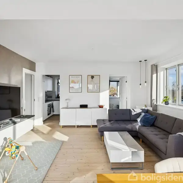 Stue med grå sofa og sofabord, hvor tv'et er slukket. Lyst træ som gulvbelægning. Store vinduer giver dagslys. Legetøj på gulvet, malerier på væggen og adgang til køkkenet.