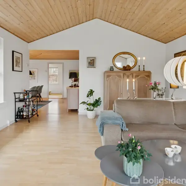 Stue med hjørnesofa i brun, træbord med planter og lysestager, lyse trægulve og loft. Vinduer langs væggen til venstre, åbning til køkkenområde med spiseplads i baggrunden.