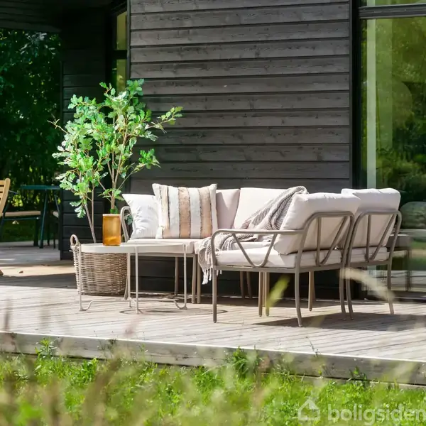 Sofa står på en træterrasse foran et mørkt træhus, omgivet af grønne planter og en anden træstol. Puder er placeret på sofaen, skaber en hyggelig atmosfære.