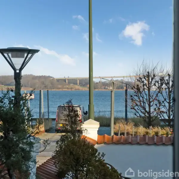 En have ses gennem en døråbning. Træer og buske står stille. I baggrunden krydser en bro over en havnefront på en solrig dag.