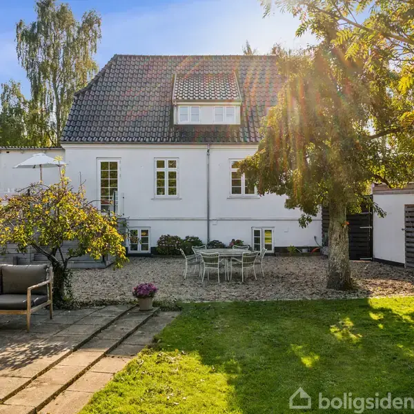 En hvid murstensbygning med sadeltag står i en solrig have. En grusgårdsplads med havemøbler er omgivet af træer og grønne plæner.