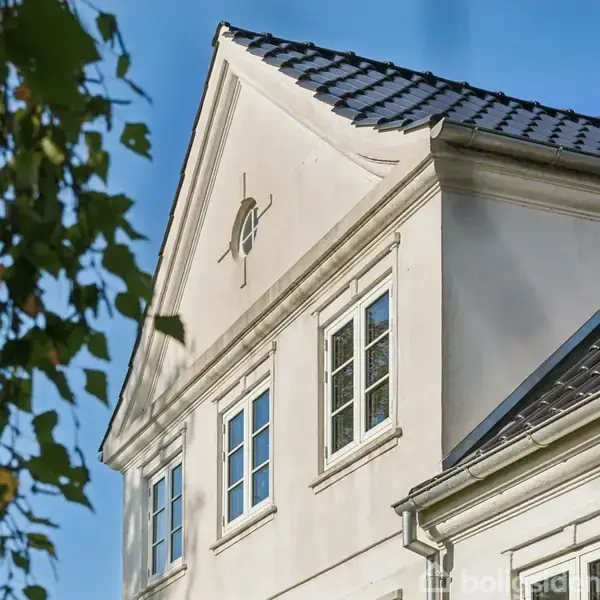 Et hvidt hus med sort tegltag står under en blå himmel. Trægrene med grønne blade ses i forgrunden, som rammer det øverste hjørne af huset ind.