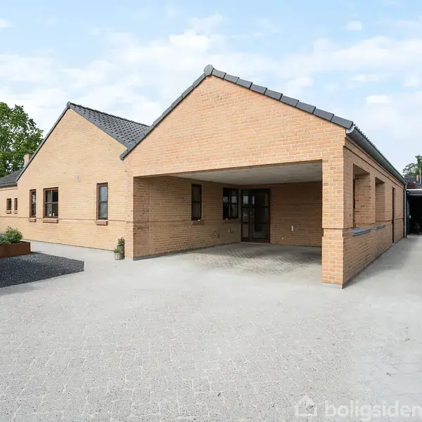 Et moderne hus i røde mursten med carport, beliggende langs en flisebelagt indkørsel. Omkranset af grønne buske og træer, ses et klar himmel i baggrunden.