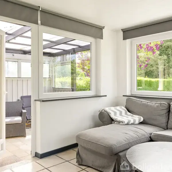 Grå sofa står i en lys stue med store vinduer, der åbner op til en overdækket terrasse. Udsigt til grøn have udenfor.