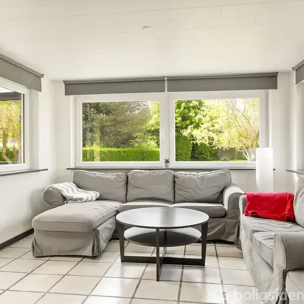 Grå hjørnesofa med rødt tæppe, placeret i lys stue med hvide vægge og klinkegulv. Store vinduer viser grøn natur udenfor. Rundt sofabord står foran sofaen.