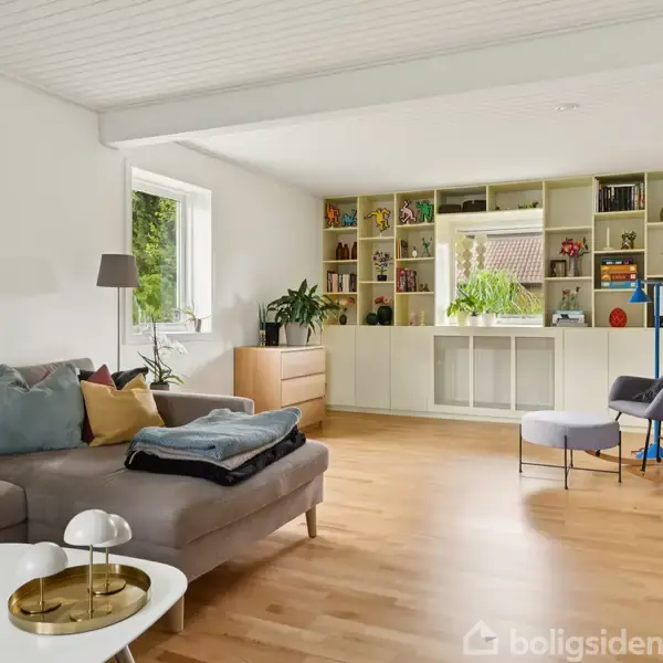 Stue med grå sofa, sofabord med lampe, og reol med bøger og planter. Vinduer giver lys, og trængerammer giver farve. Trægulv og hvide vægge skaber en lys og moderne atmosfære.