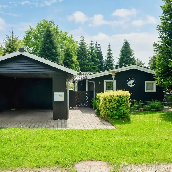 Sort træhus med carport til venstre, omgivet af grønne træer og en velplejet græsplæne, under en klar blå himmel. Verandaen har en dekorativ cirkel med silhuet af en fisk.