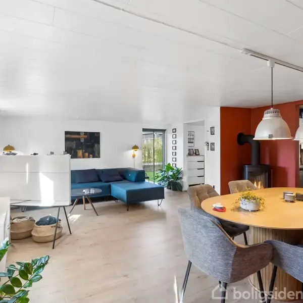 Spisebord med stole står i lys stue, omgivet af planter, en blå sofa og dekorative lamper. Store vinduer giver klart lys, og åbningen til et køkken ses i baggrunden.