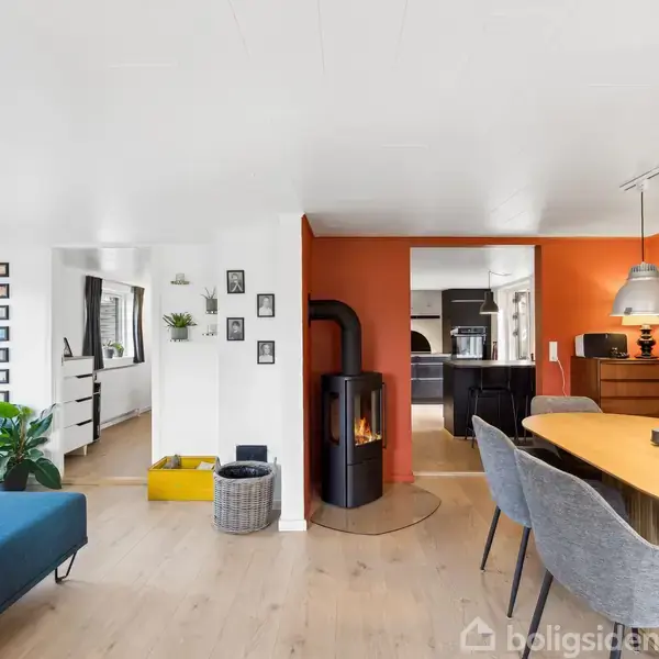 Ovn på væggen brænder ved siden af et rundt spisebord med grå stole; placeret i en lys stue med blå sofa og grønne planter. Flot loftbelysning og orangerøde vægge.