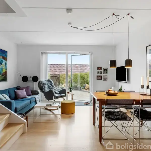 En moderne stue med en blå sofa og et træbord. Sofaen står til venstre ved siden af vinduet, og bordet er omgivet af stole. Væggene har kunstværker.