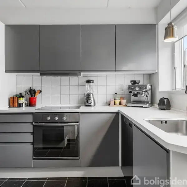 Gråt køkken med indbyggede skabe. På bordpladen står en blender og espresso-maskine. Et stort vindue med udsigt giver naturligt lys. En vask er integreret i bordpladen.