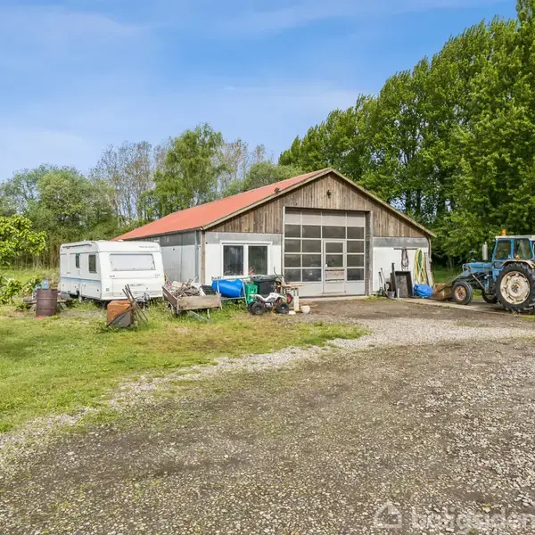 En stor garage med rødt tag står ved en grusvej. En traktor og en campingvogn ses foran, omgivet af grønne træer og græsarealer.