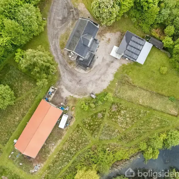 Hus med grusvej omgivet af træer i frodig grøn natur. Rød længebygning ligger ved siden af. Vandkanten er synlig i nederste højre hjørne.