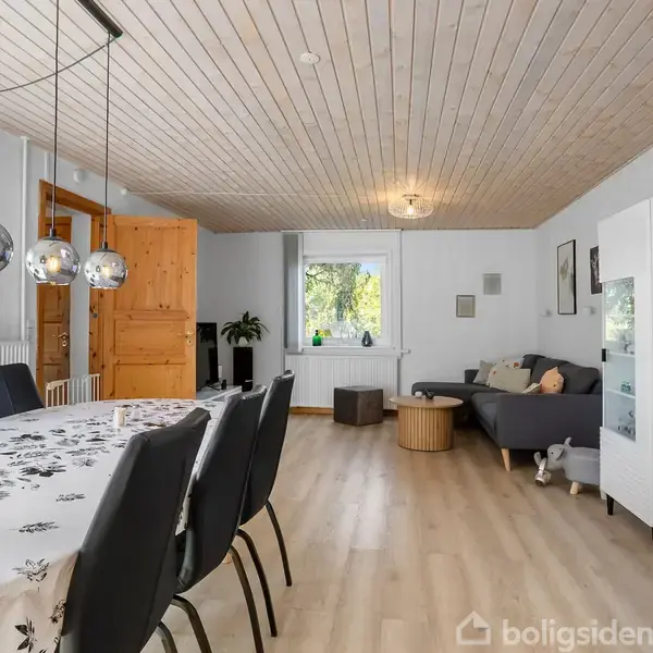 Spisestue med sort spisebord og seks stole, dækket med hvid dug. I baggrunden er en grå sofa og rektangulært vindue. Trædør til venstre, træloft, tre runde lamper hænger over bordet.