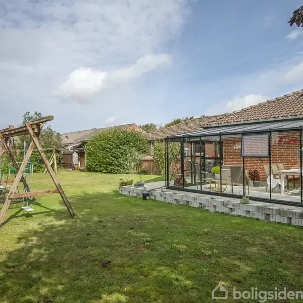 Et gyngestativ står på en græsplæne ved siden af et hus med en overdækket terrasse. En blå himmel og enkelte skyer ses i baggrunden.