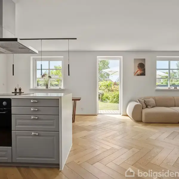 Køkkenområde med gråt skab og indbygget ovn, åbner ind til stue med beige sofa og træbord. Store vinduer og dør leder ud til en grøn have.