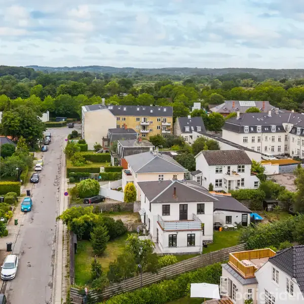 Boligområde med hvide og farverige hustage omgivet af grønne haver og træer. Biler parkerer langs vejen. Baggrund viser skovklædte bakker under en skyet himmel.