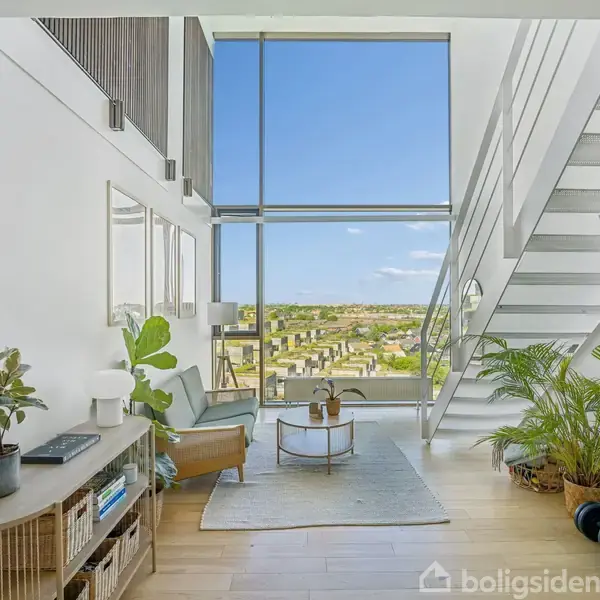 En moderne stue med store vinduer vender mod en byudsigt. En sofa og et bord står på et tæppe. Planter og en reol med bøger skaber en hyggelig atmosfære.