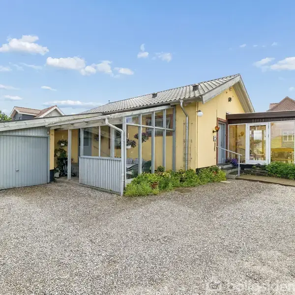 Et gult hus med tagterrasse har glasindgang og tilknyttet garage. Det står i en have med grusgang. I baggrunden ses andre huse og en blå himmel med få skyer.