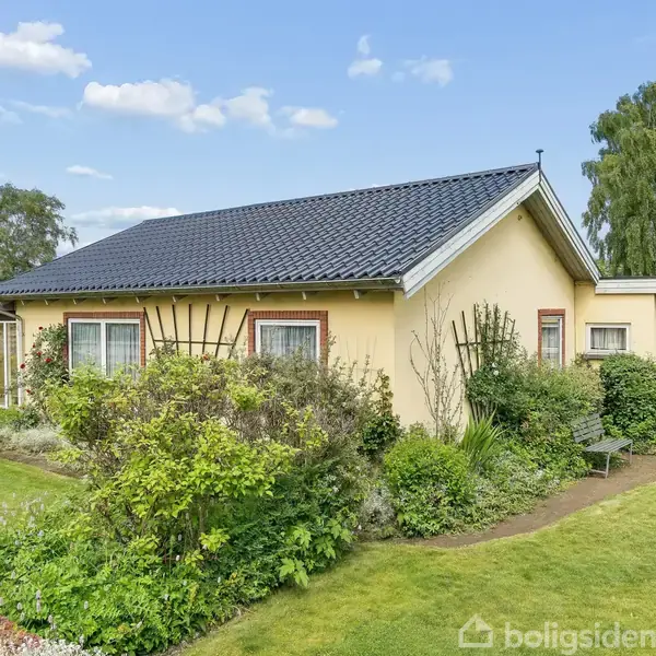 Et gulmalet hus med sort tag står omgivet af en velplejet have med buske og træer. Himlen er blå med få skyer i baggrunden.
