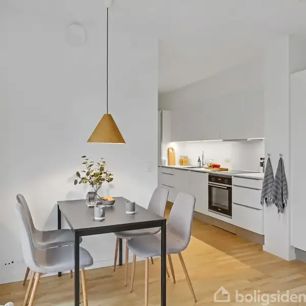 Spisebord med stole står under en pendellampe i køkken-alrum. Baggrunden viser et hvidt, moderne køkken med indbygget ovn og skabe. En plante og stearinlys pynter bordet.