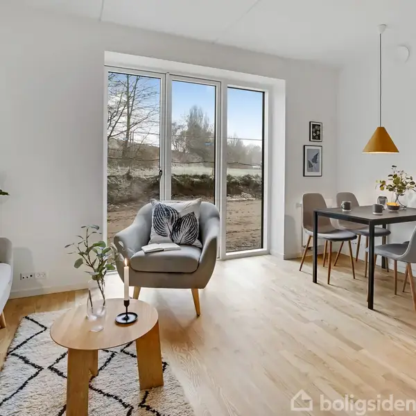 Stue med en lænestol ved et stort vindue, omkranset af en sofa og et sofabord. I baggrunden ses et spisebord med stole og en hængende lampe.