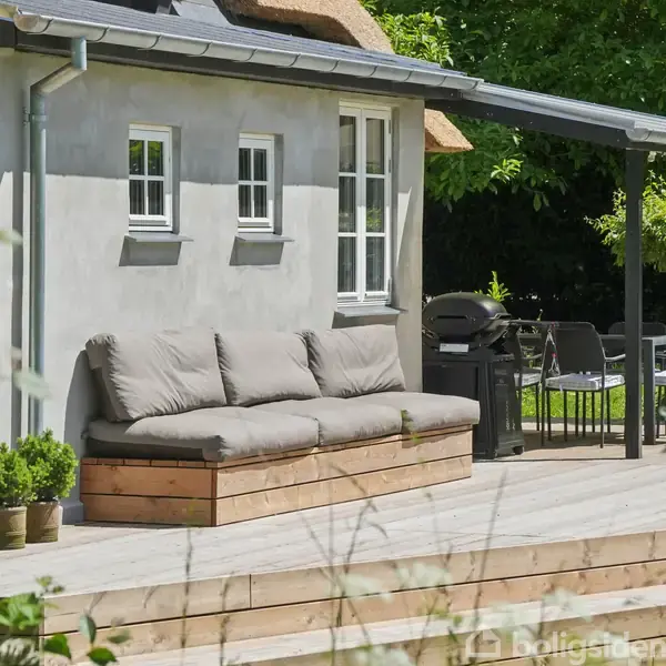 Terrasse med en grå sofa og puder står foran et hus med hvid facade. Havebord og stole ses i baggrunden, omkranset af grønne buske og træer.