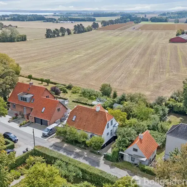 Flere huse med røde tage står langs en vej. Bag dem strækker store, åbne marker sig ud til horisonten. Træer omkranser området, der fremstår grønt og landligt.